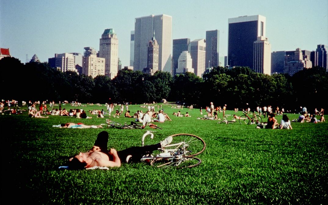 Film still from CENTRAL PARK. People are lying in the grass in Central Park, in the background you can see the skyscrapers of Manhattan.
