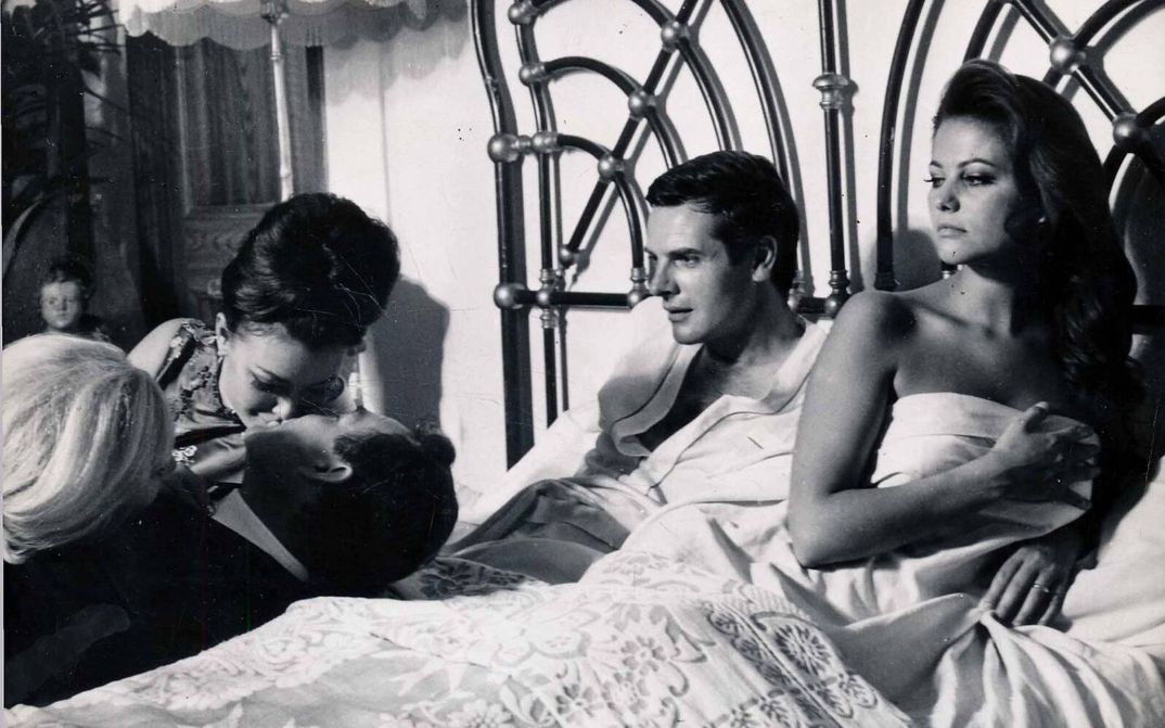 Film still from IL MAGNIFICO CORNUTO: A man and a woman are lying in bed. They look at three people beside the bed, two of whom are kissing.