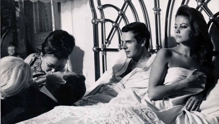 Film still from IL MAGNIFICO CORNUTO: A man and a woman are lying in bed. They look at three people beside the bed, two of whom are kissing.