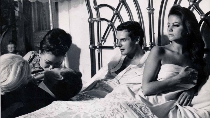 Fimstill aus IL MAGNIFICO CORNUTO: Ein Mann und eine Frau liegen im Bett. Sie blicken auf drei Personen neben dem Bett, von denen zwei sich küssen.