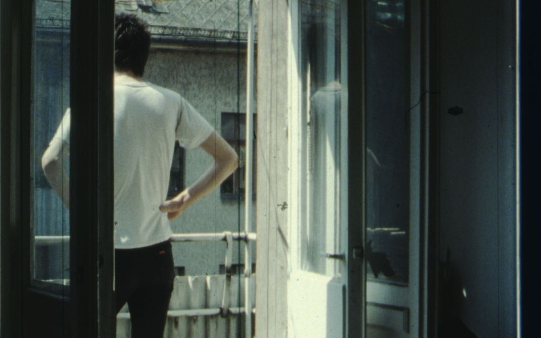 Filmstill aus IM SOMMER: Eine Person steht auf dem Balkon, die Hände in die Hüfte gestützt.