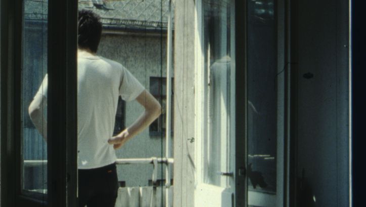 Filmstill aus IM SOMMER: Eine Person steht auf dem Balkon, die Hände in die Hüfte gestützt.