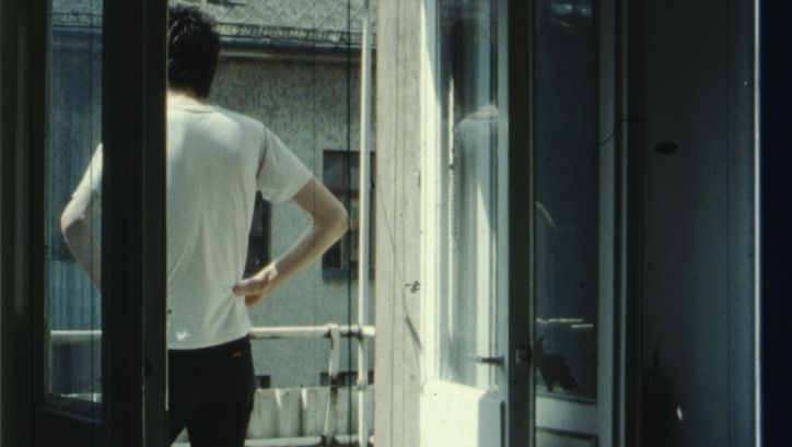 Filmstill aus IM SOMMER: Eine Person steht auf dem Balkon, die Hände in die Hüfte gestützt.