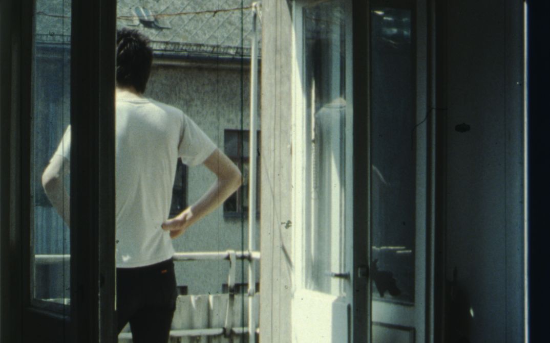 Filmstill aus IM SOMMER: Eine Person steht auf dem Balkon, die Hände in die Hüfte gestützt.
