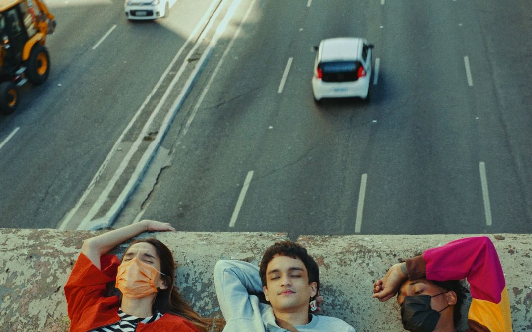 Film still from TRES TIGRES TRISTES: You see the heads of three young people on a wall. Behind them, cars can be seen on a road.
