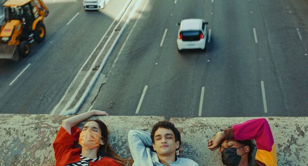 Filmstill aus TRES TIGRES TRISTES: Man sieht die Köpfe dreier junger Menschen auf einer Mauer. Hinter ihnen sind Autos auf einer Straße zu sehen.
