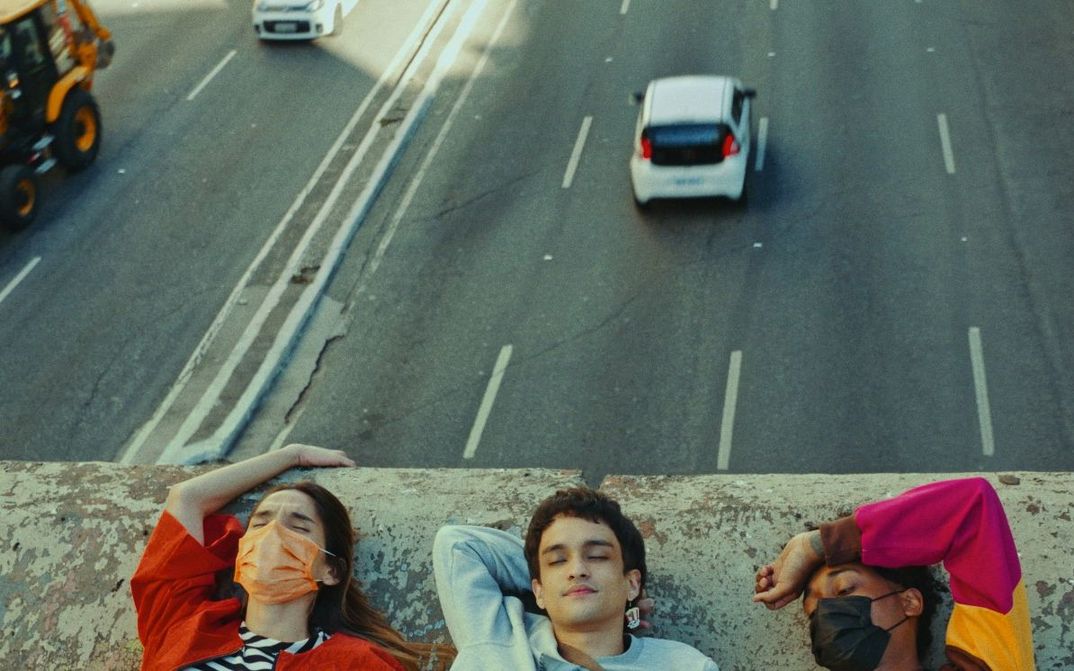 Film still from TRES TIGRES TRISTES: You see the heads of three young people on a wall. Behind them, cars can be seen on a road.