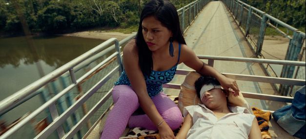 Film still from PUNKU by J.D. Fernández Molero. The picture shows a woman sitting on a narrow bridge. In her arms is a blindfolded person.