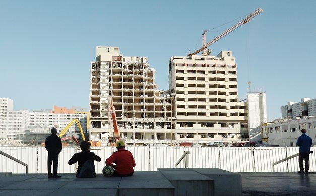 Film still from BERLIN JWD: A few people watch as a high-rise building is demolished.