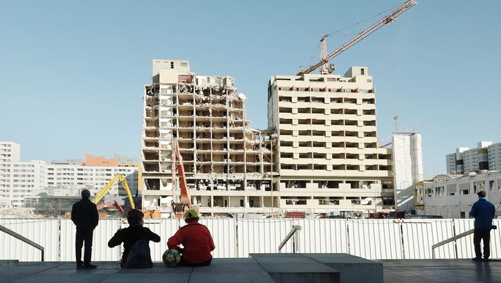 Film still from BERLIN JWD: A few people watch as a high-rise building is demolished.