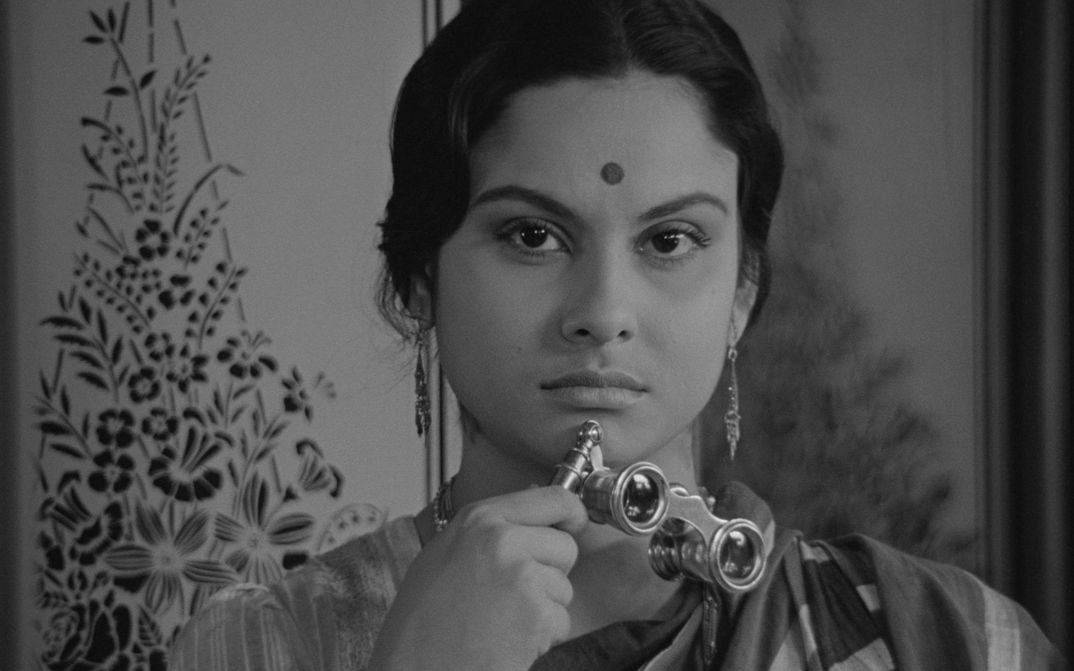 Filmstill from CHARULATA: A woman looks into the camera. She has a dot on her forehead and is holding opera glasses.