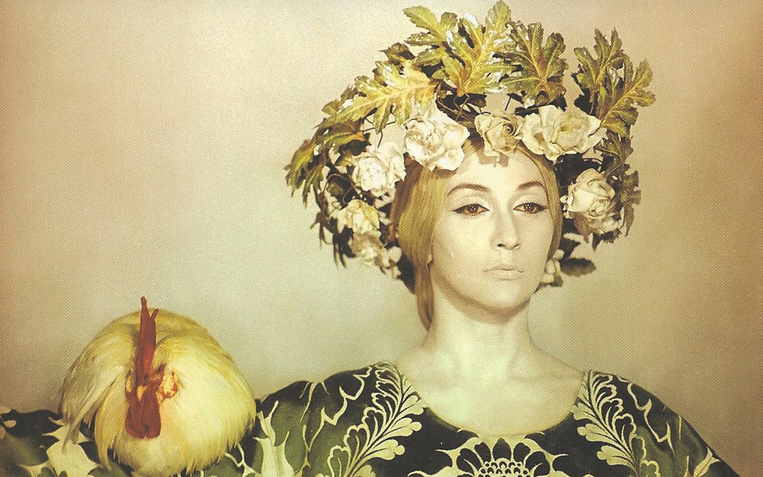 Film still from SAYAT NOVA: A yellowish colored picture with a woman with striking makeup and hair ornaments, on her shoulder sits a chicken.