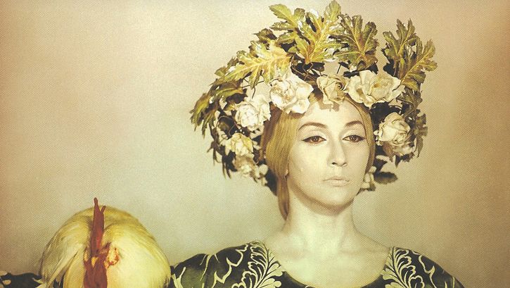 Film still from SAYAT NOVA: A yellowish colored picture with a woman with striking makeup and hair ornaments, on her shoulder sits a chicken.