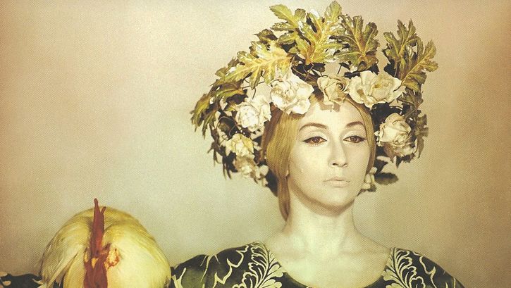 Film still from SAYAT NOVA: A yellowish colored picture with a woman with striking makeup and hair ornaments, on her shoulder sits a chicken.