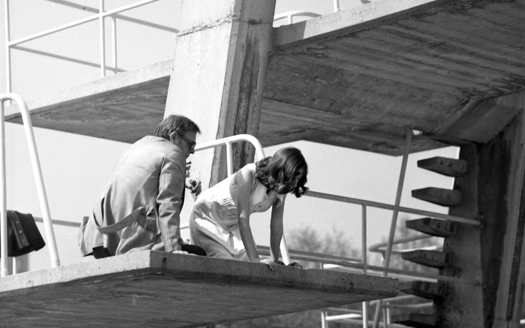 Film still from THE SWIMMING POOL. A woman and a man, both dressed, are sitting on the diving tower of an outdoor swimming pool.