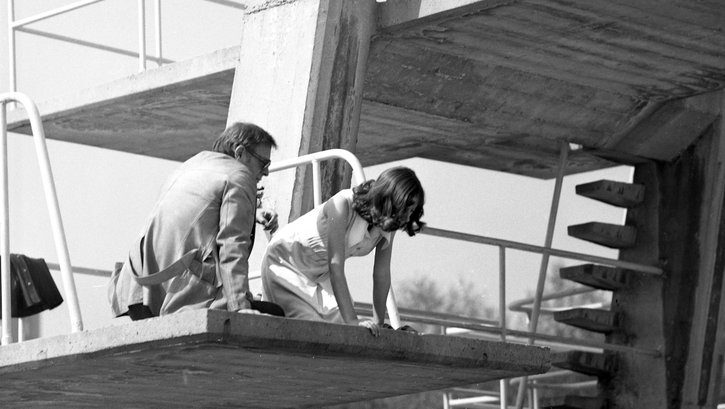 Film still from THE SWIMMING POOL. A woman and a man, both dressed, are sitting on the diving tower of an outdoor swimming pool.