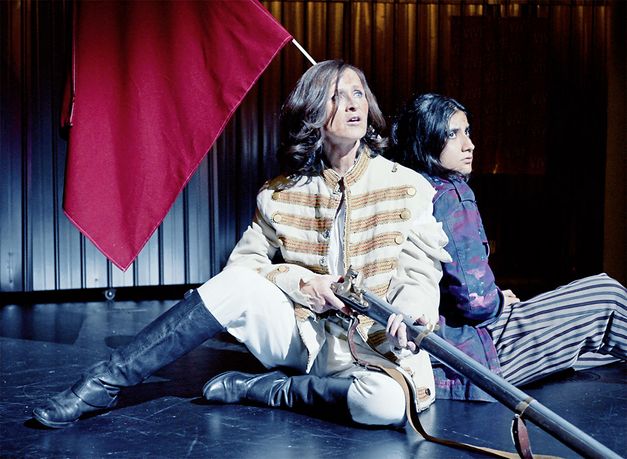 Still from the film "L’état et moi" by Max Linz. Two women in fantasy uniforms sit on a stage. One holds an old-fashioned rifle in her hand. 