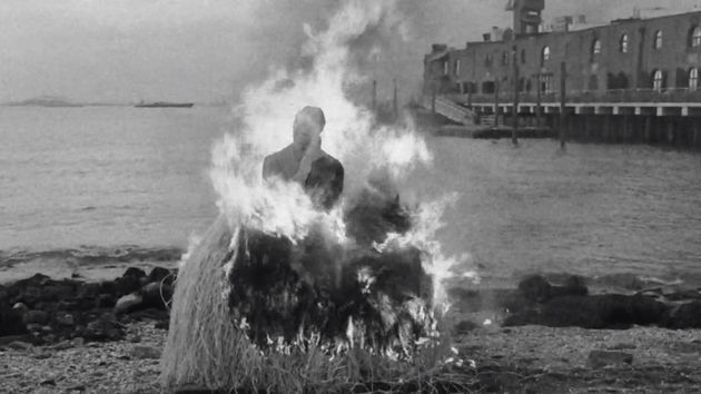 Filmstill aus dem Film „Conspiracy“ von Simone Leigh, Madeleine Hunt-Ehrlich. Ein menschliches Bildnis, welches vor einer Wasserfläche in einem großen Feuer verbrennt.