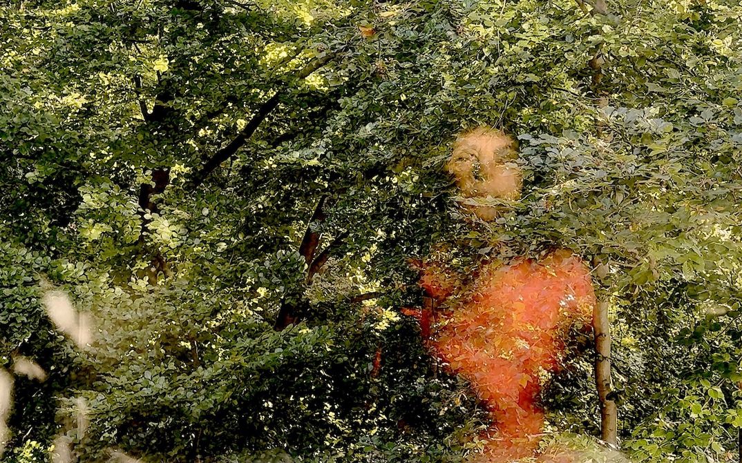 Filmstill aus HOPE AGAINST HOPE: Man sieht dicht Blätter von Bäumen, verschwommen ist das Bild einer Frau im roten Pulli hineinmontiert.