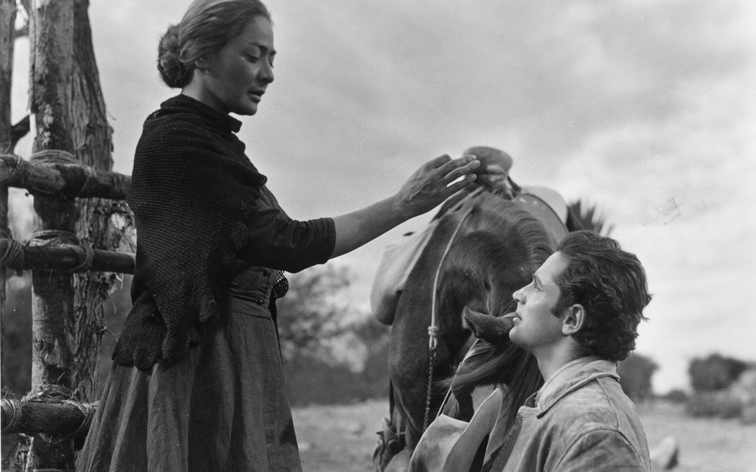 Film still from LOS HERMANOS DEL HIERRO: A woman bends down to a man standing next to a horse.