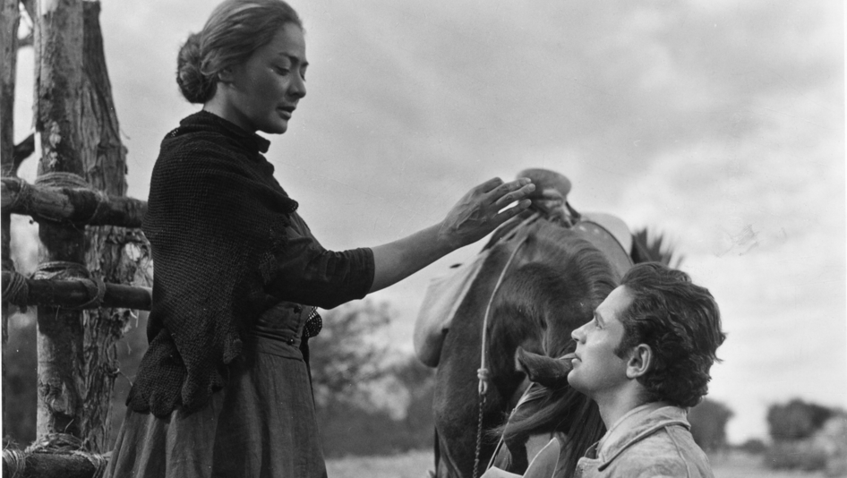 Film still from LOS HERMANOS DEL HIERRO: A woman bends down to a man standing next to a horse.