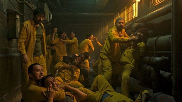 Eine Gruppe Männer in gelber Kleidung kämpfen miteinander in einem engen, dunklen Gang mit Rohren an den Wänden.