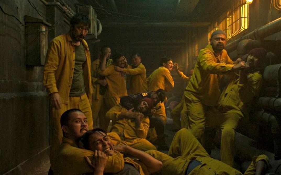 Eine Gruppe Männer in gelber Kleidung kämpfen miteinander in einem engen, dunklen Gang mit Rohren an den Wänden.