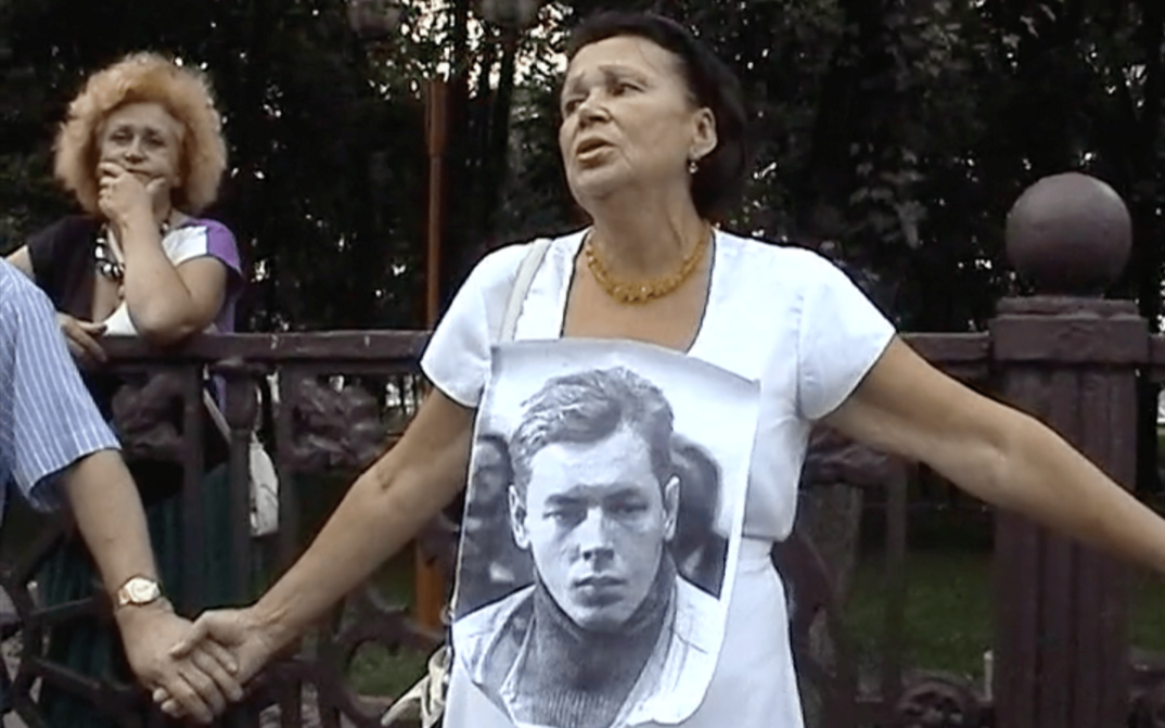 Film still from I WAS CALLED TO THE BALL: A few women hold hands during a protest. The woman in the center wears a photo of a young man around her neck.
