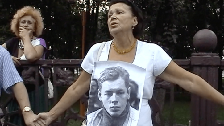 Film still from I WAS CALLED TO THE BALL: A few women hold hands during a protest. The woman in the center wears a photo of a young man around her neck.