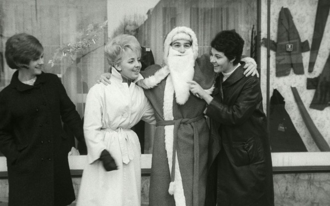 Film still from LE PÈRE NOEL A LES YEUX BLEUS: Three women are standing in front of a store window next to Santa Claus, joking with him.