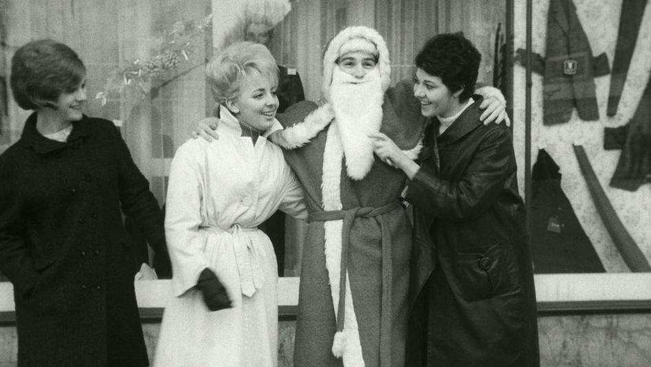 Film still from LE PÈRE NOEL A LES YEUX BLEUS: Three women are standing in front of a store window next to Santa Claus, joking with him.
