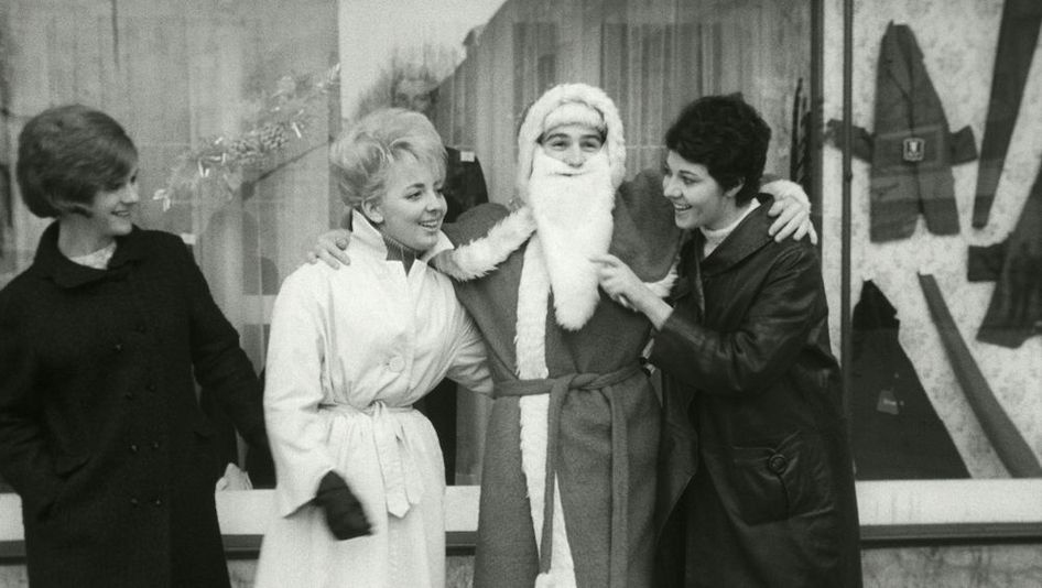 Film still from LE PÈRE NOEL A LES YEUX BLEUS: Three women are standing in front of a store window next to Santa Claus, joking with him.