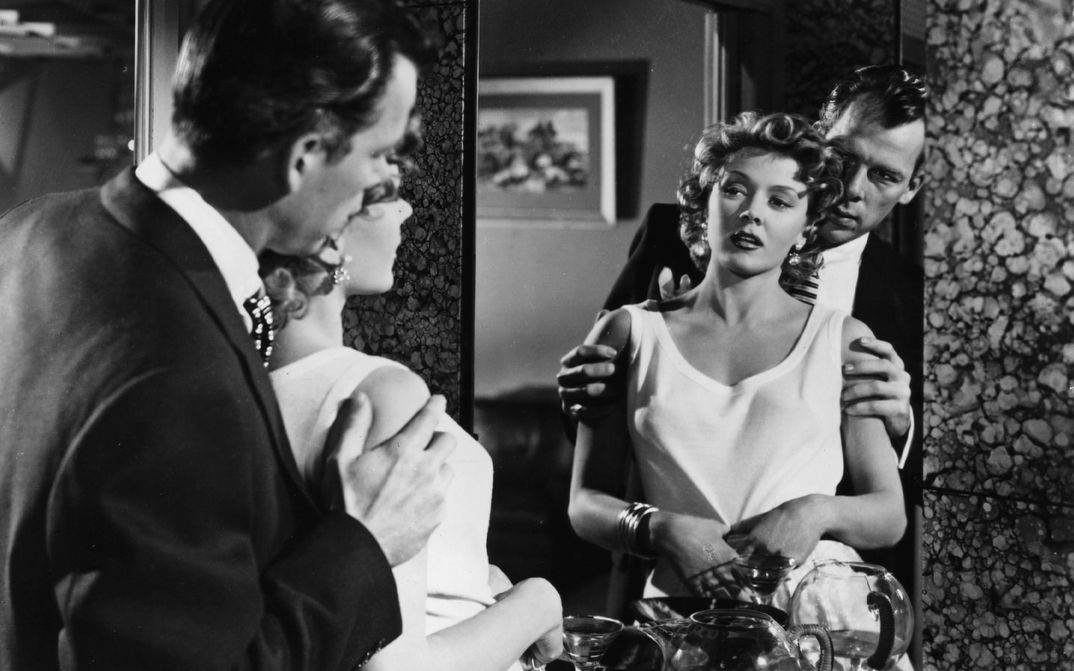 Filmstill aus THE BIG HEAT: Ein Mann und eine Frau betrachten sich im Spiegel. Er steht hinter ihr und hält sie an den Armen.