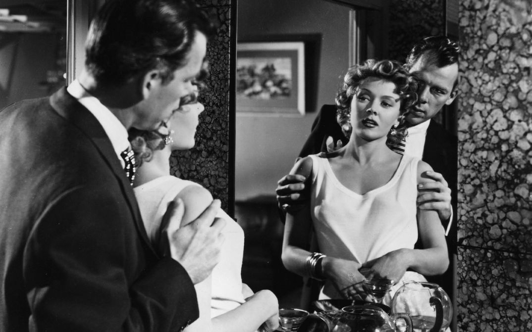 Film still from THE BIG HEAT: A man and a woman look at themselves in the mirror. He stands behind her and holds her by the arms.