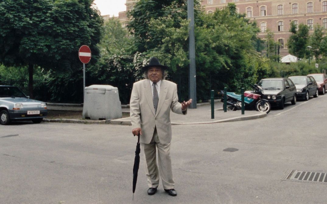 Filmstill aus WANKOSTÄTTN: Ein Mann in grauem Anzug, Krawatte und schwarzem Zylinder stützt sich auf einen Regenschirm. Er steht auf der Straße. Im Hintergrund sind geparkte Autos, Bäume und Häuser zu sehen.