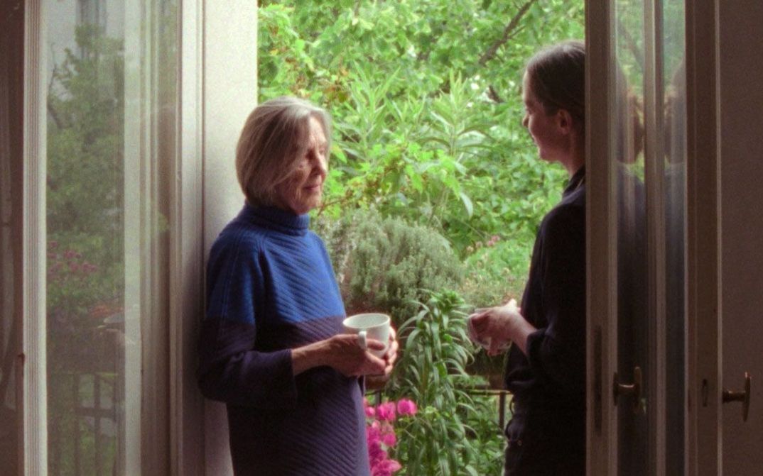 Two women stand in an open balcony doorway, one holding a cup, talking in front of lush plants.