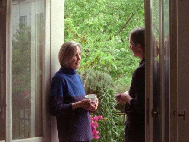 Zwei Frauen stehen in einer geöffneten Balkontür, eine hält eine Tasse, beide im Gespräch vor üppiger Bepflanzung.
