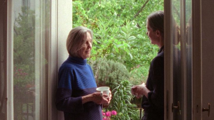 Two women stand in an open balcony doorway, one holding a cup, talking in front of lush plants.