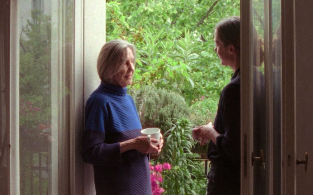 Two women stand in an open balcony doorway, one holding a cup, talking in front of lush plants.