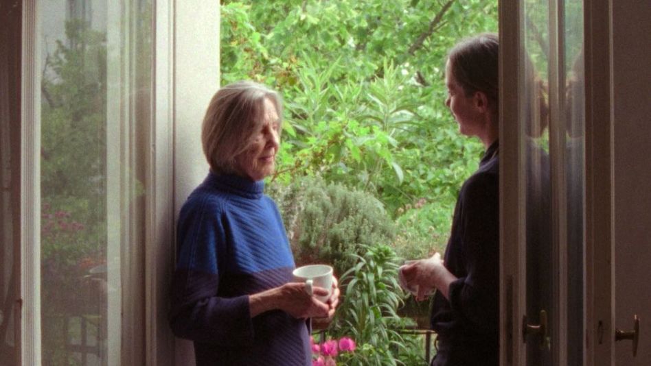 Two women stand in an open balcony doorway, one holding a cup, talking in front of lush plants.