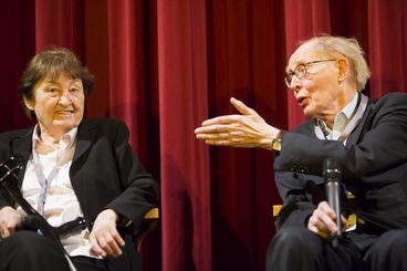 The two are sitting in front of a red curtain, speaking.