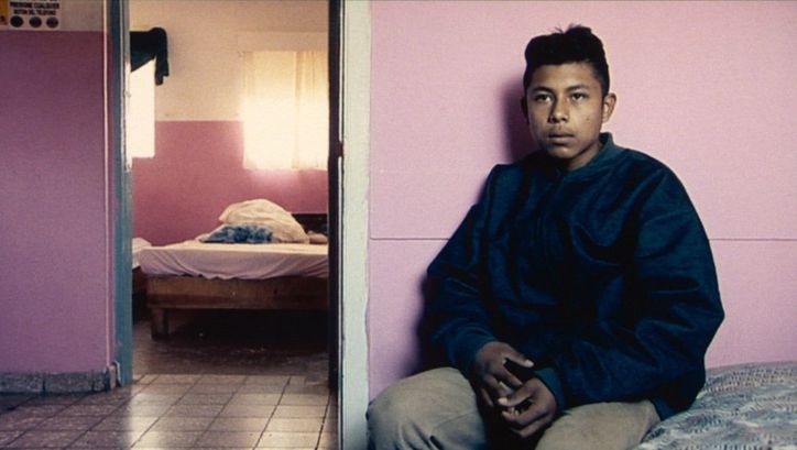 Film still from DE L’AUTRE CÔTÉ: A young man sits on a bed in sparse accommodation and looks into the camera.