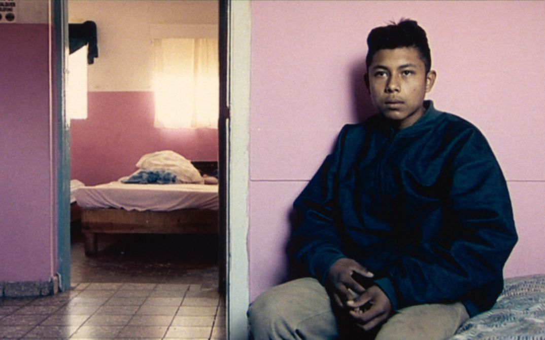 Film still from DE L’AUTRE CÔTÉ: A young man sits on a bed in sparse accommodation and looks into the camera.