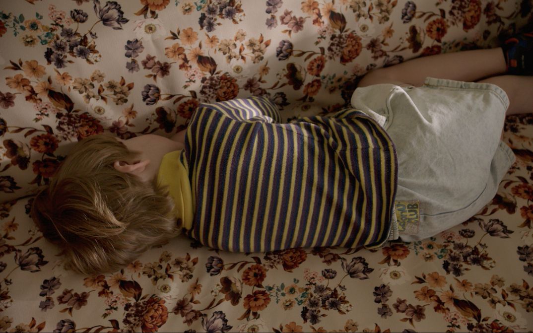 Film still from THE CATHEDRAL. A child is lying on a sofa with a floral pattern, with his back facing the viewer.