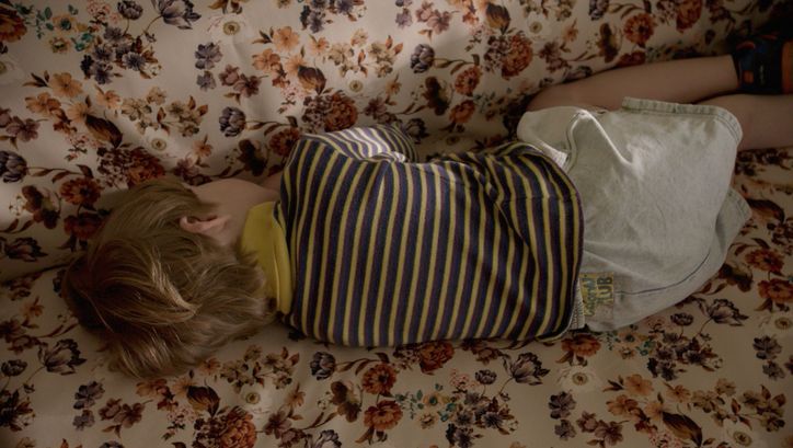 Film still from THE CATHEDRAL. A child is lying on a sofa with a floral pattern, with his back facing the viewer.