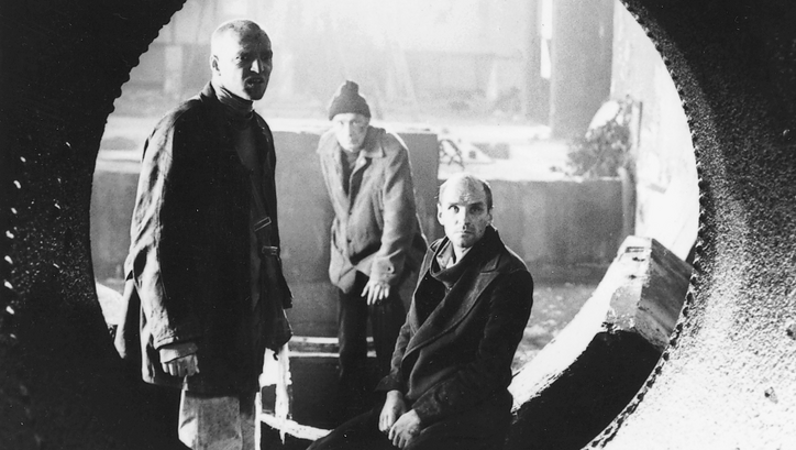 Film still from STALKER: Through a pipe, you can see three men standing in an industrial hall.