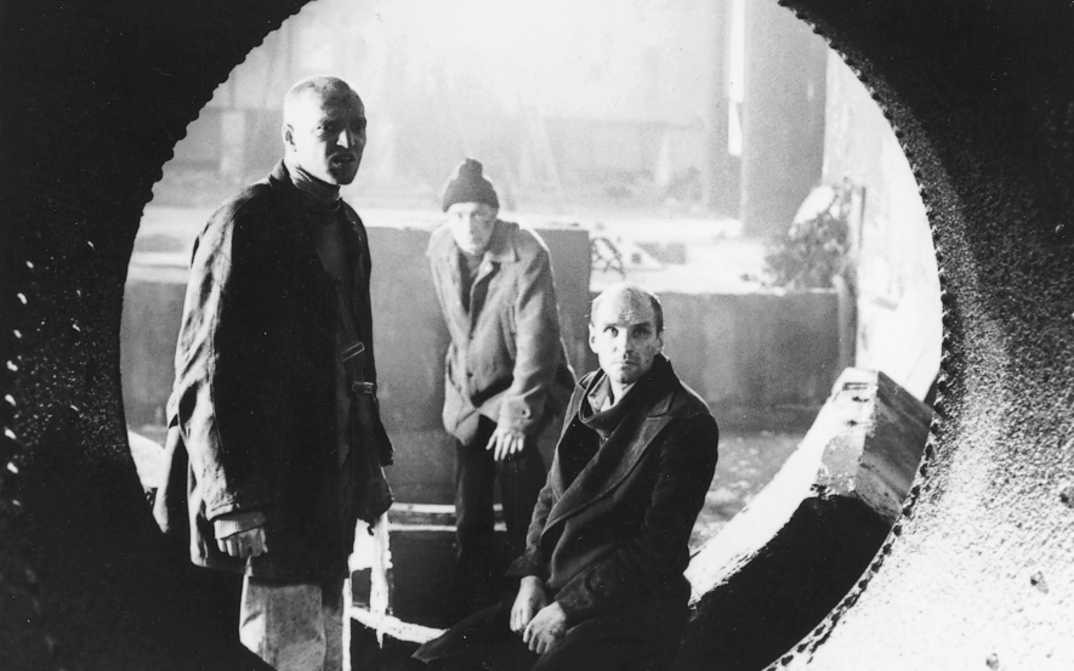 Film still from STALKER: Through a pipe, you can see three men standing in an industrial hall.