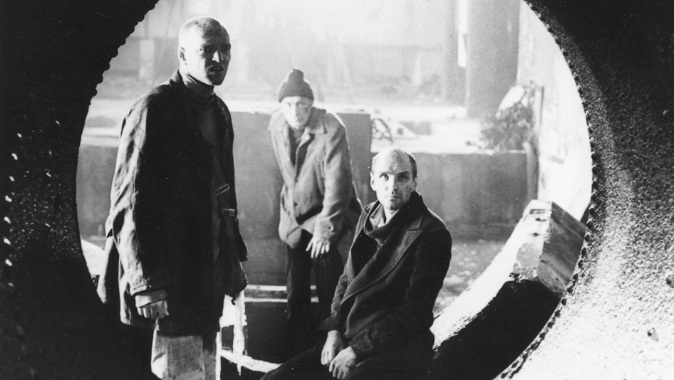 Filmstill aus STALKER: Durch ein Rohr blickt man auf drei Männer, die in einer Industriehalle stehen.