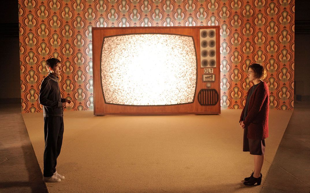 It shows a stage that gets wider towards the back. A large television is placed at the back wall in the middle of the picture. On the left there is a man and on the right a woman who are looking at each other and talking.