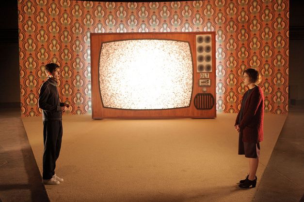 It shows a stage that gets wider towards the back. A large television is placed at the back wall in the middle of the picture. On the left there is a man and on the right a woman who are looking at each other and talking.